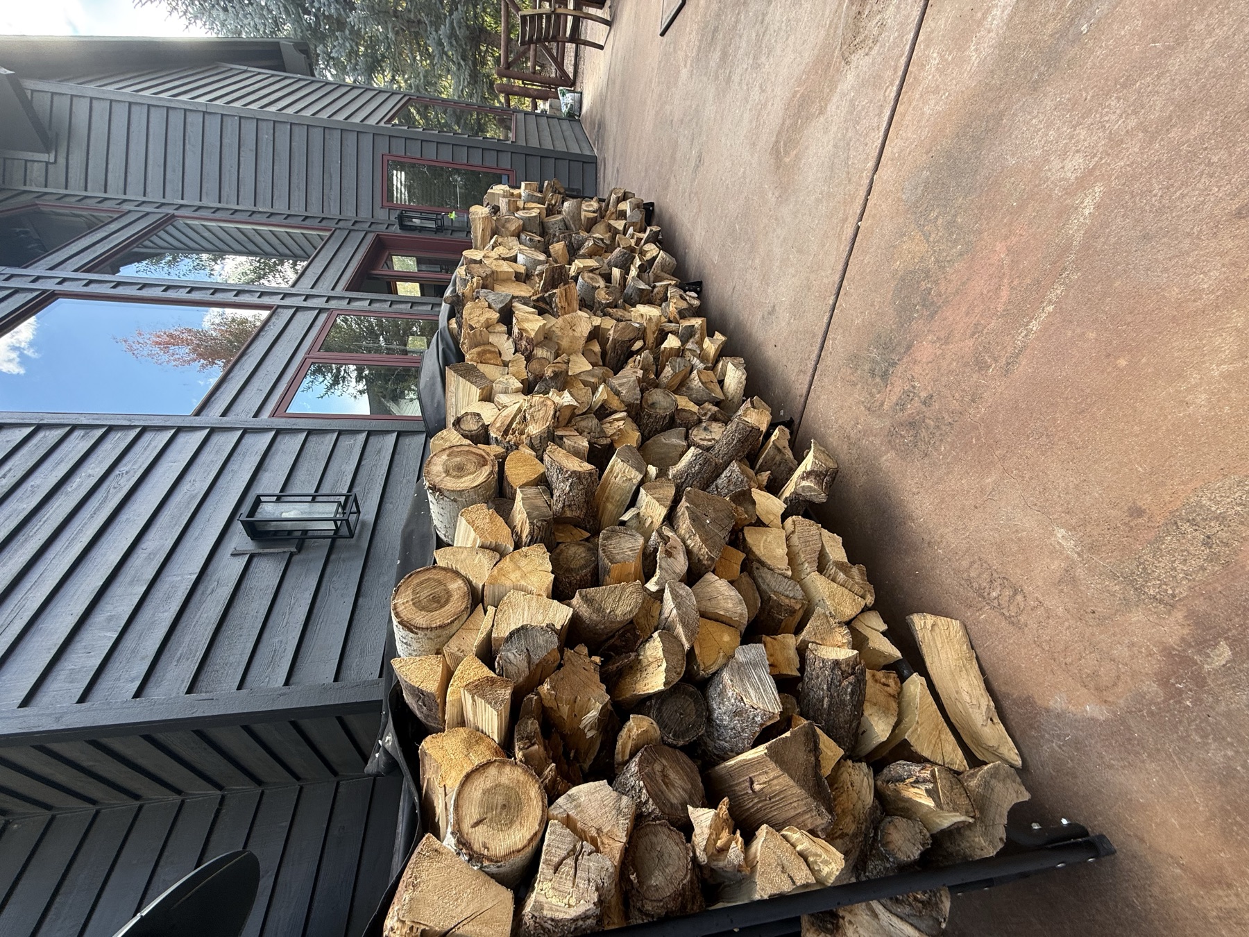 Stacked firewood on a steel rack beside a patio, split ends facing out, mountain home behind